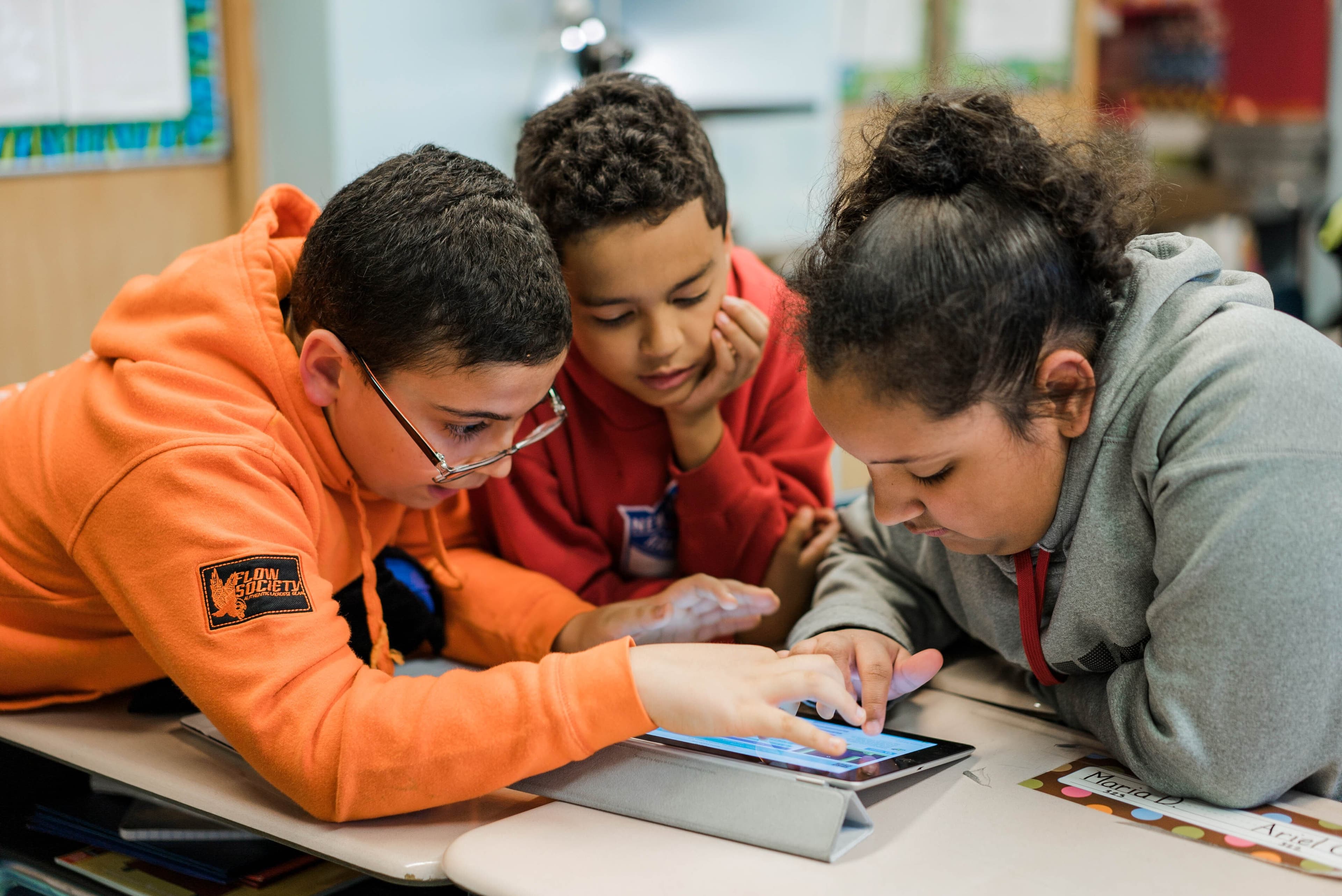 Three students leaning in together over a tablet in a classroom