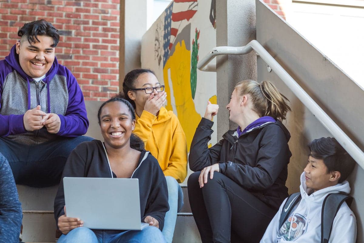 A group of smiling students sitting together on a school staircase, one with a laptop open